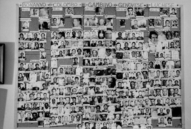 Large bulletin board showing photographs of bosses, underbosses, capos and soldiers in five NY organized crime families, Bonanno, Colombo, Gambino, Genovese and Luchese. 