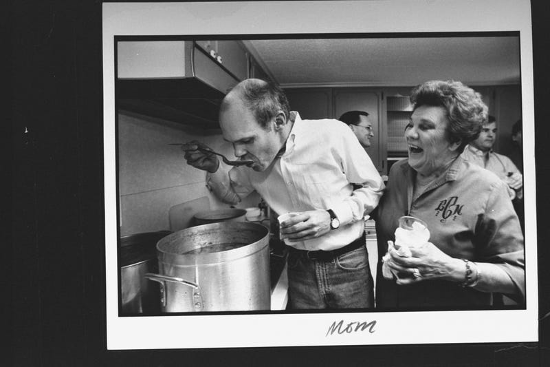 James Carville, tasting a spoonful of soup from large pot on stove while his mom Lucille laughs while looking on in her kitchen at home.