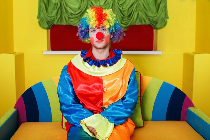 Clown in rainbow colored costume with makeup and red nose sitting on colorful sofa