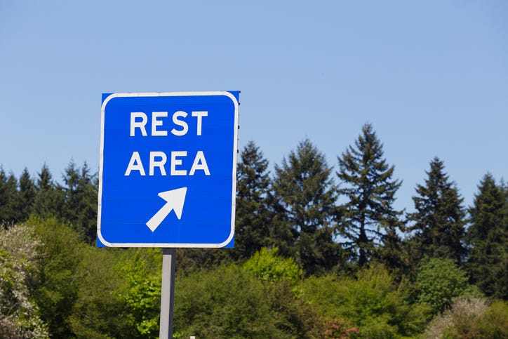 Close up shot of a rest area sign on the side of a highway. A wooded area with evergreens and other trees can be seen in the background.