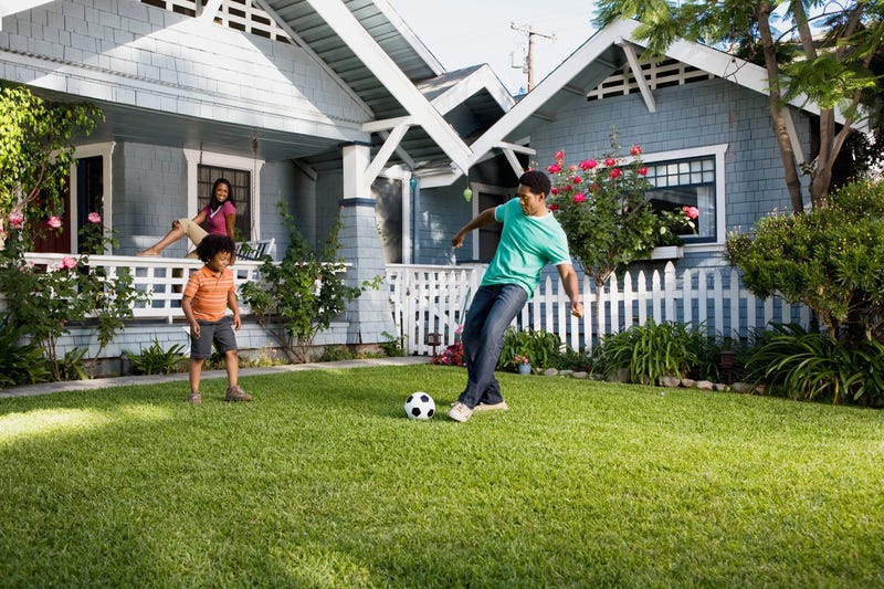 A father and son kick a soccer ball in a yard while a mother looks on