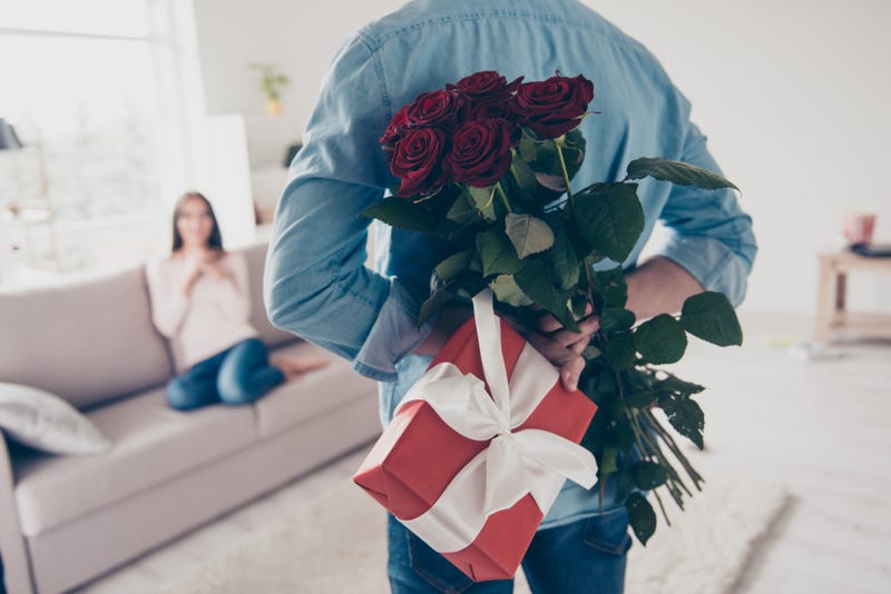 stock image. woman sits on couch, man stands in front of camera facing her holding roses and a gift behind his back for valentine's day