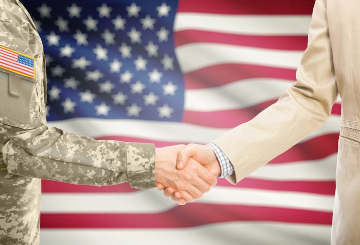 USA military man in uniform and civilian man in suit shaking hands with American flag in background