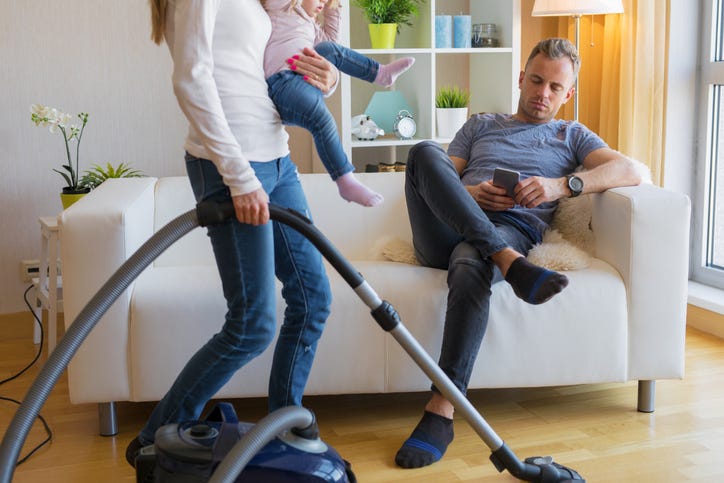 Woman Vacuums While Man Sits With His Phone