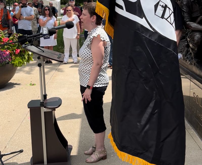 Woman sings at podium under a POW-MIA flag
