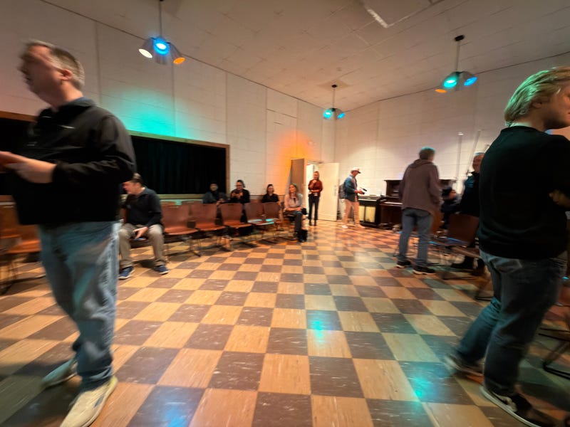 A group of people in a retro checkered-tile room with ambient orange and blue lighting. Some are seated while others stand and walk, creating a relaxed atmosphere.
