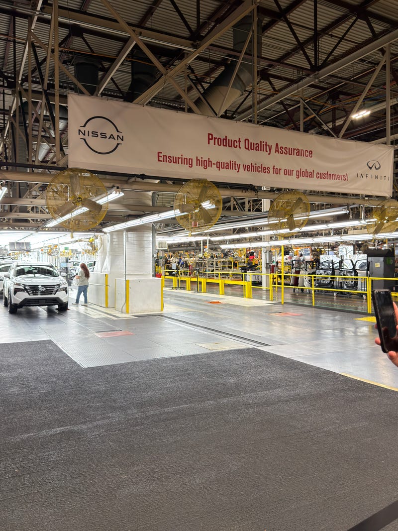 Car manufacturing plant with a Nissan banner reading "Product Quality Assurance." A worker stands near a white car. Industrial setting, efficient atmosphere.