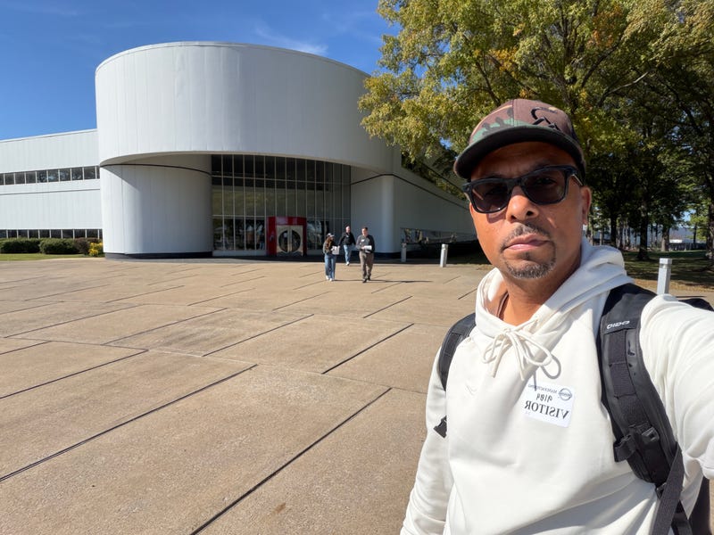 A man in sunglasses and a hoodie takes a selfie outside a modern, curved building on a sunny day. Three people walk in the background.