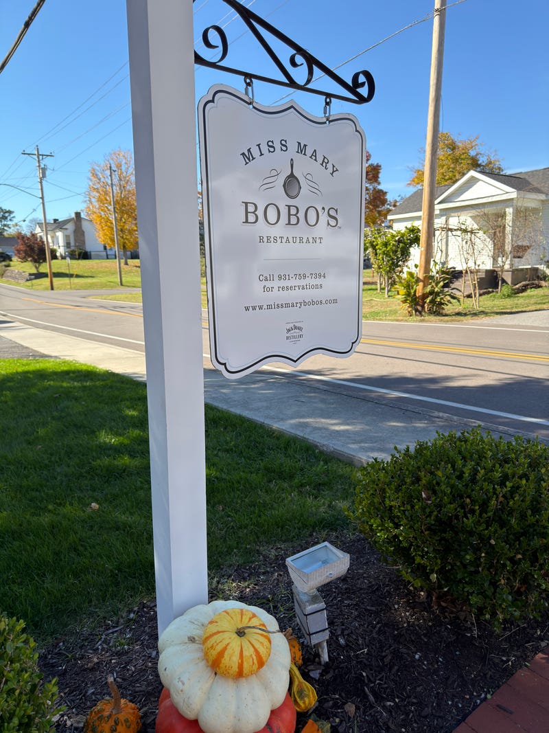 Outdoor sign for "Miss Mary Bobo's Restaurant" hangs next to a road on a sunny day, surrounded by autumnal foliage and decorative pumpkins.