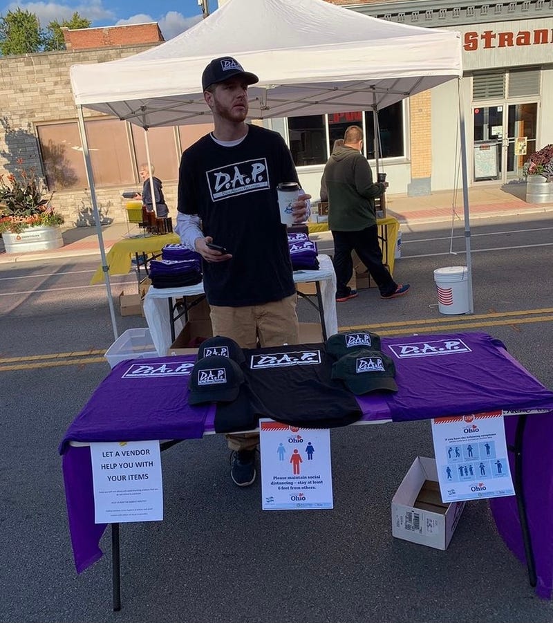 A man wearing a D.A.P t-shirt standing behind table of D.A.P. merchandise