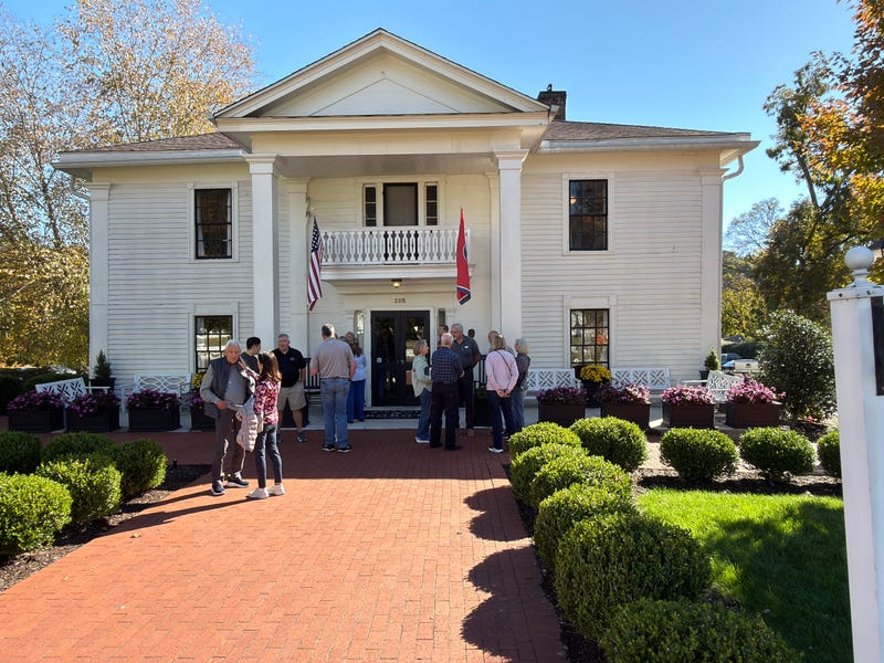 Historic white building with columns and two flags, surrounded by lush greenery and flowers. A group of people is gathered outside on a sunny day.