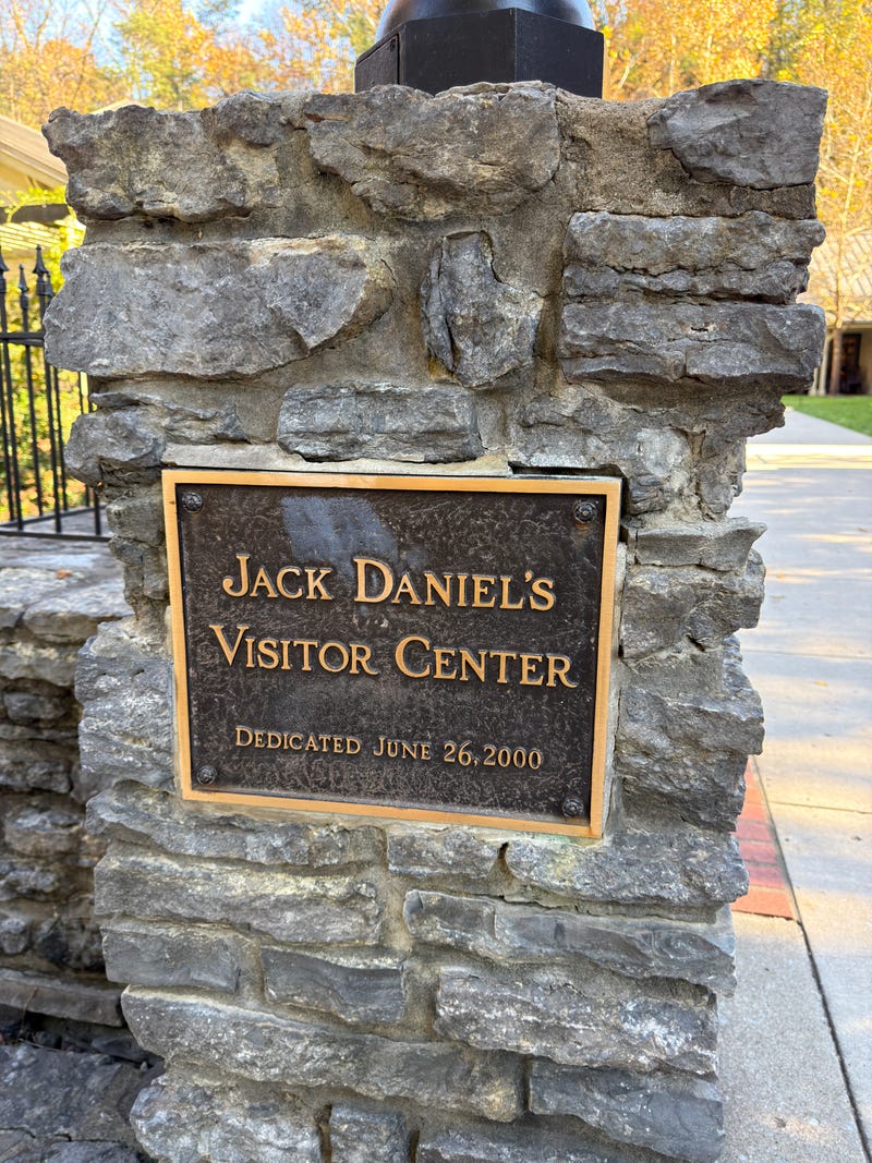 Plaque on a stone post reads "Jack Daniel's Visitor Center, Dedicated June 26, 2000." Surrounded by fall foliage and a paved walkway.