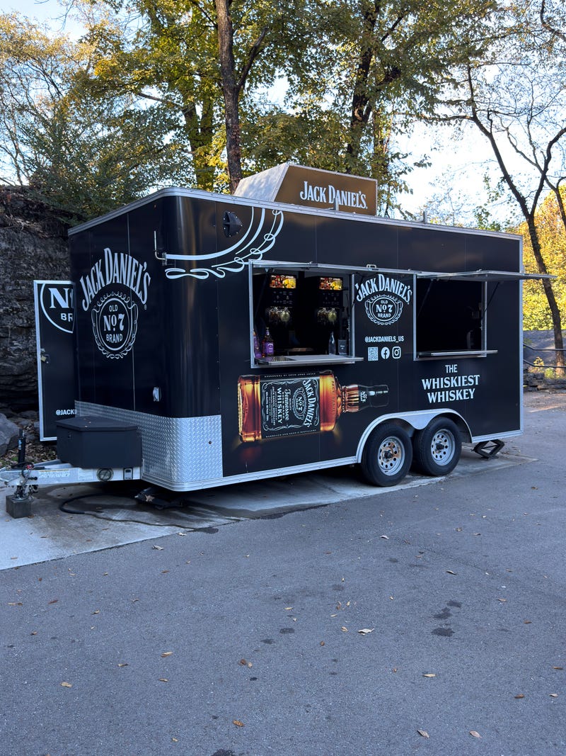 Mobile whiskey bar set in a serene outdoor scene with trees. The black trailer features Jack Daniel's branding and a prominent whiskey bottle image.