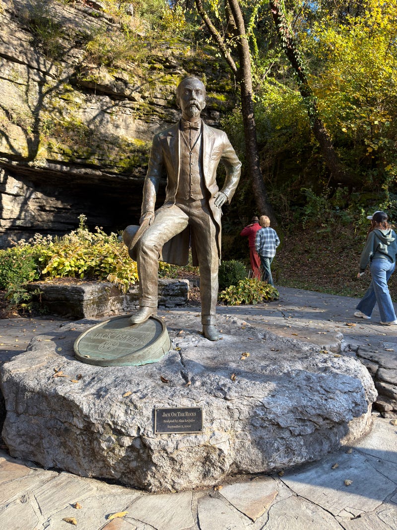 A bronze statue of a man standing on a large rock, set against a natural backdrop of trees and a rocky hill. Two people walk by, and sunlight filters through leaves.