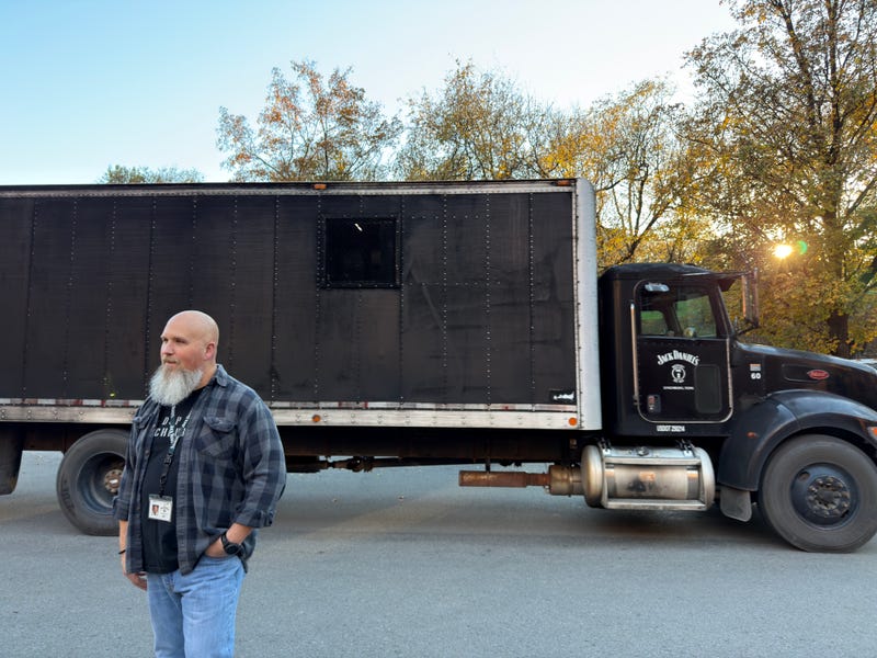 A bearded man stands in front of a large black truck with "Jack Daniels" branding. Trees with autumn leaves are in the background, with a setting sun.