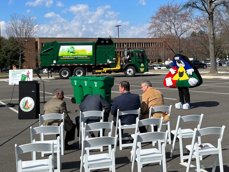 A colorful recycling-themed mascot stands beside 3 super cans and an empty lecturn.  Henrico representatives look on.
