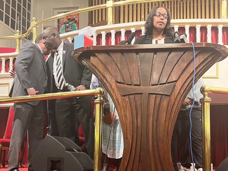 A woman stands at a lectern, speaking. Several men and women stand behind her.