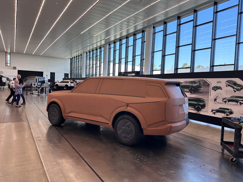 Clay SUV model in a well-lit design studio with large windows. Concept sketches on the wall. People in background convey a busy, creative atmosphere.