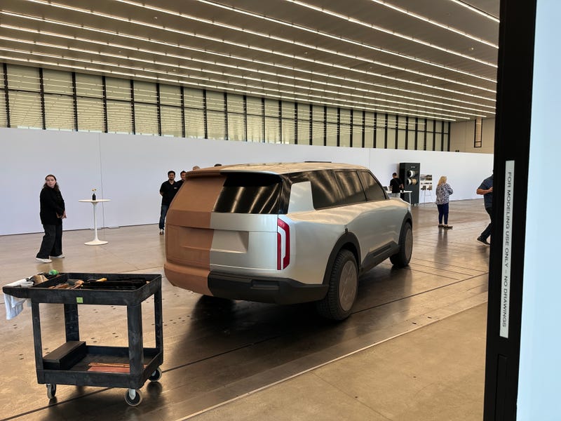 Clay model of a futuristic SUV in a large, well-lit design studio. People observe the model, suggesting a focus on innovation and creativity.
