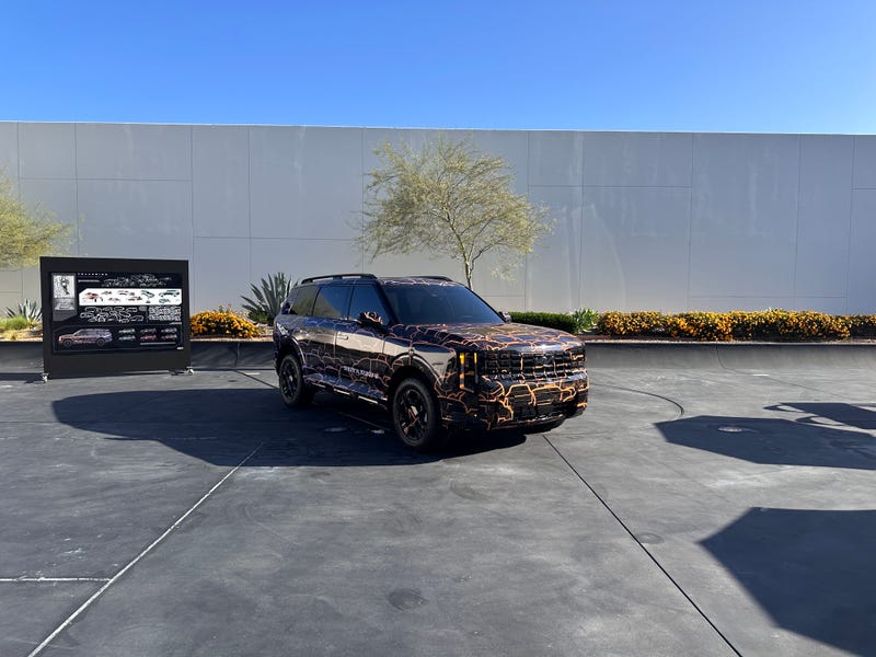 SUV with a striking black and orange camouflage pattern parked on a sunny concrete lot, near a display board and surrounded by plants and a modern wall.