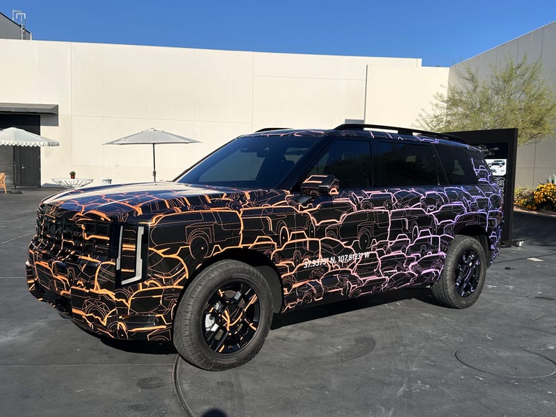 SUV with a striking black and orange camouflage pattern parked on a sunny concrete lot, near a display board and surrounded by plants and a modern wall.