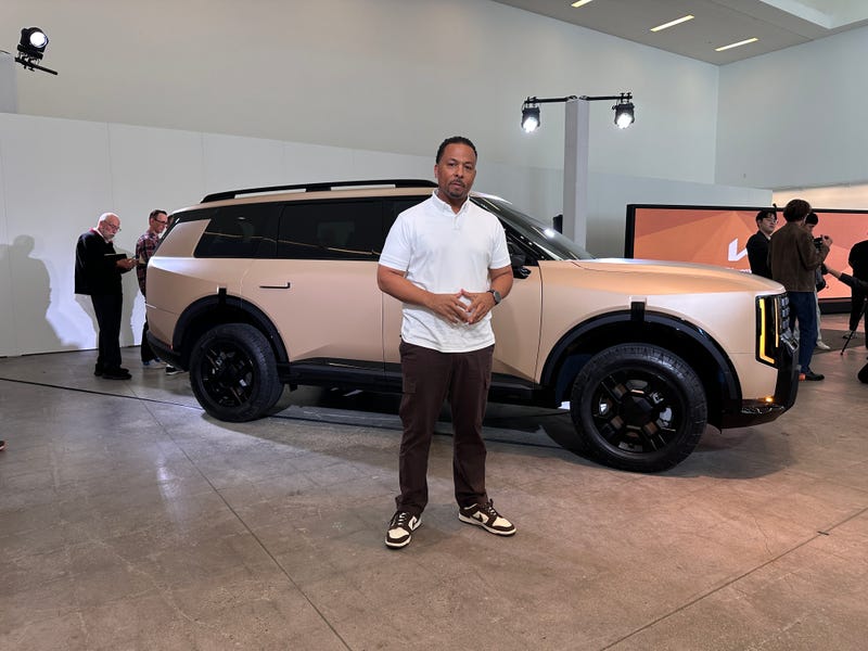 A man in a white shirt stands confidently in front of a sleek, tan SUV displayed indoors. The room has bright lighting, with a casual, modern atmosphere.