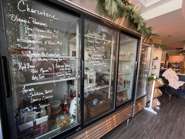 A giant open kitchen overlooks cozy wooden tables and tan, puffy pillows underneath soft lit chandeliers. There is retail from local artists and cheeses and charcuterie from Illinois growers in front.