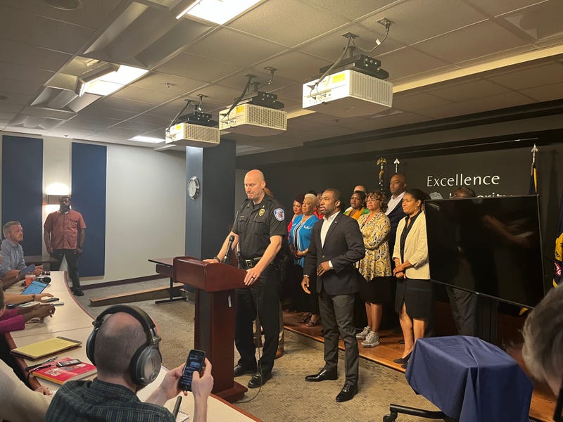 Police chief, with the Mayor, the Superintendent, and a small crowd of local leaders, addresses a room full of reporters
