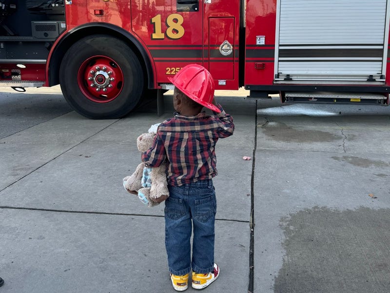 Jayveon with teddy bear and firefighter hat in front of fire truck