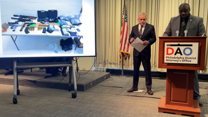 Assistant DA Keion Cook speaks at a press conference next to a photo displaying guns, cash and weapons found in the investigation into a West Philly drug ring.