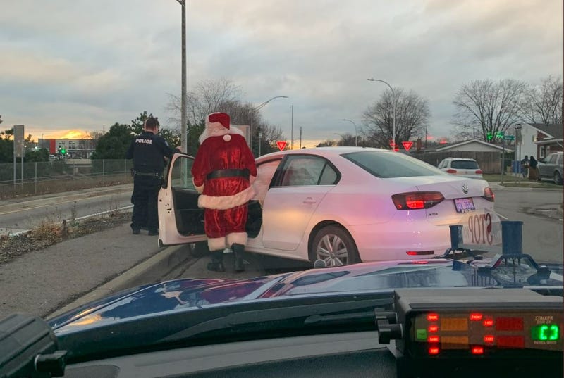 Picture taken from the dash of a police car. A white Volkswagen sedan is crashed into a curb. The driver is dressed as Santa Claus. He's standing just outside the car, speaking with a Michigan State Police Trooper.