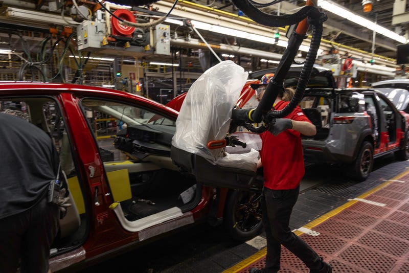 A worker in red shirt installs a car dashboard using a robotic arm on an assembly line. Red and grey cars are partially assembled, with tools overhead.
