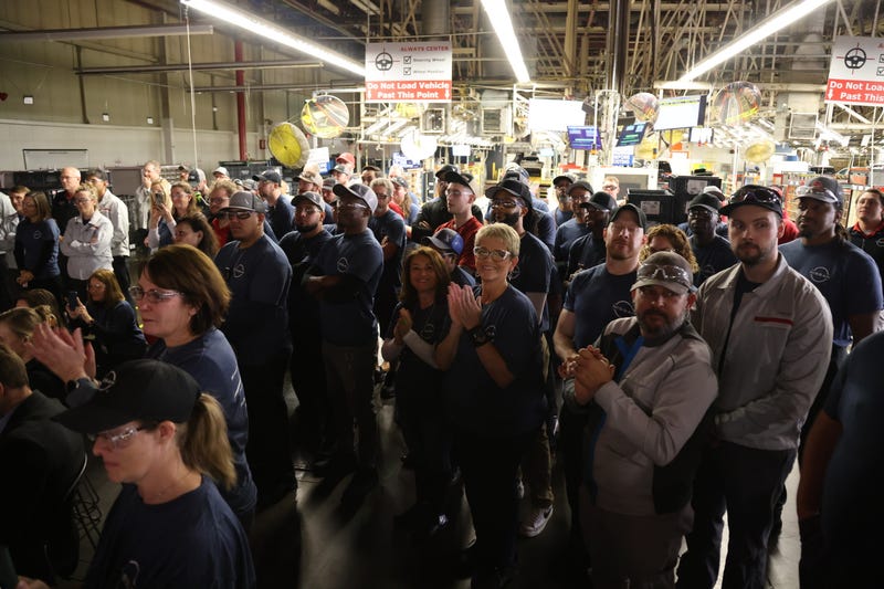 A diverse group of factory workers stands together in a brightly lit industrial space, some clapping. The mood is attentive and engaged. Signs and machinery are visible in the background.