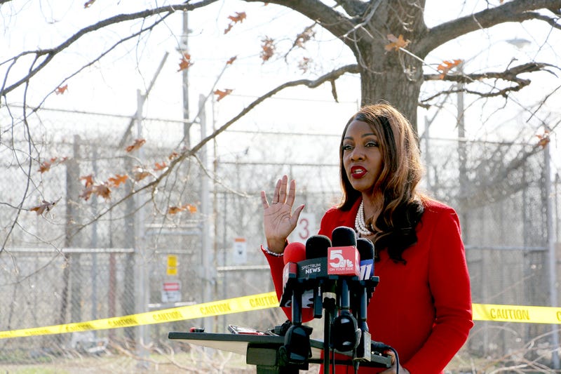 A Black woman in a red suit stands outside, in front of a fenced area. She speaks at a lectern with mics from news outlets.