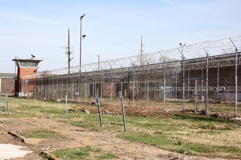 A guard tower and courtyard at a jail. Wire fences surround brick buildings in the background.