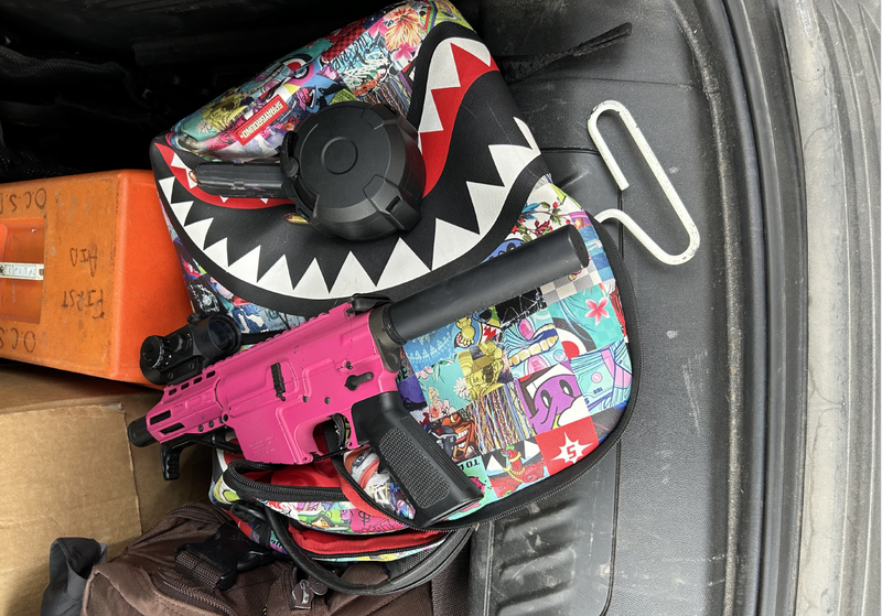 A bright, and colorful backpack featuring dozens of different pictures has a pink handgun resting on top of it.