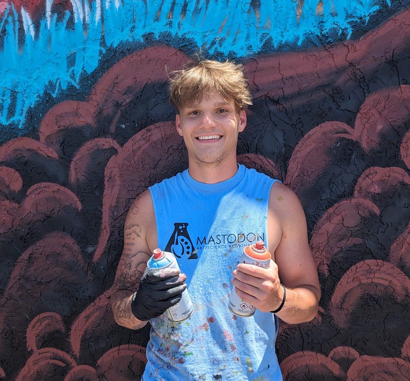 Will Breeze, 20, beginner muralist holding spray cans to complete a massive mural on Imperial Main Street.