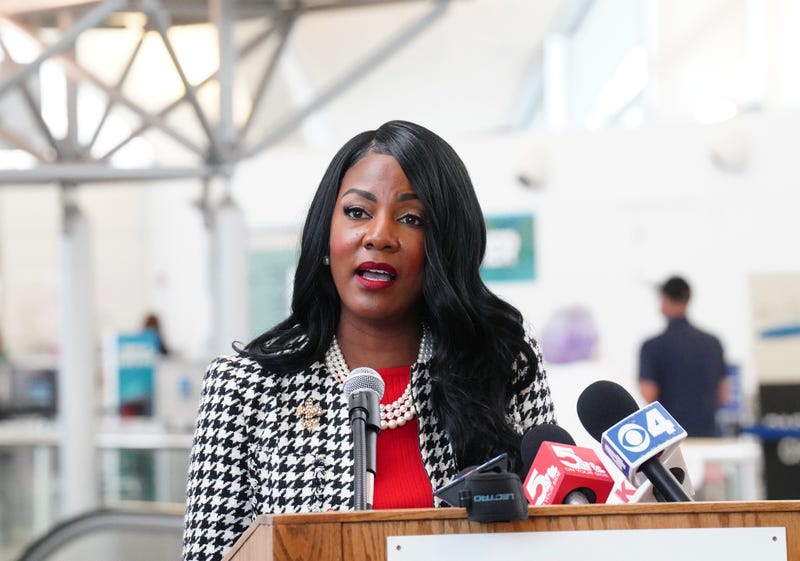 mayor jones stands at a podium speaking into mics. she wears a red shirt, a houndstooth jacket and a three-string pearl necklace.