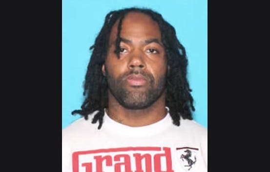 Black man in his 30s not smiling with black dreads and black facial hair wearing white t-shirt