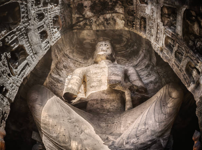 Yungang Grottoes, a UNESCO World Heritage Site in northern China. This is on a winter morning shot inside a cave where natural light filters in. There is a single huge statue in the middle with a shaft of natural light hitting it. Surrounding it on all sides are hundreds of smaller carved statues and niches. Shot using fish eye looking up to capture as much of cave as possible and create a sense of the huge scale. Image is HDR.