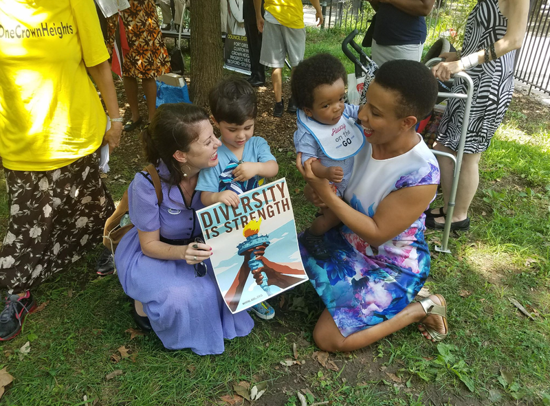 Two women, one Black and one Jewish, kneel together with young children and a sign that reads "diversity is our strength" at the One Crown Heights Neighborhood Festival.