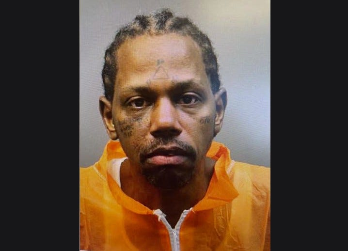 man wearing orange zip up with cornrows and face tattoos in mug shot