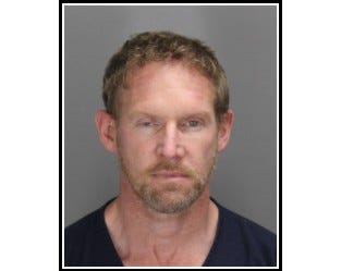 Mugshot of white man in 50s with blonde hair and beard, wearing dark v-neck