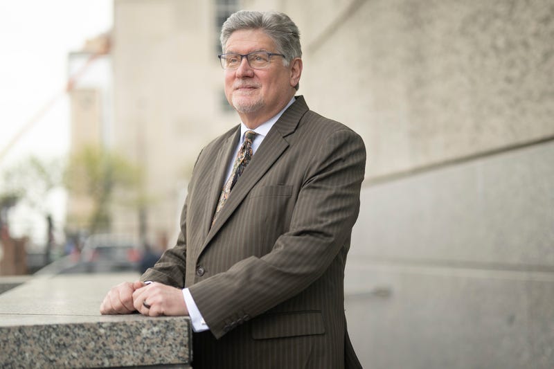 A White man wearing glasses and a tan suit poses for a portrait outside a building.
