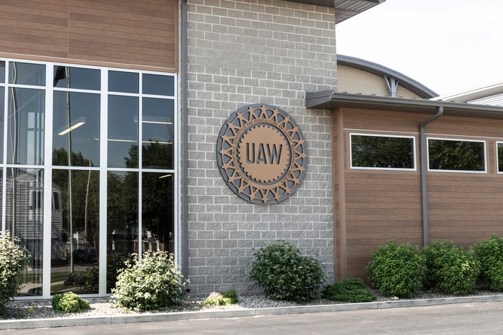 A round sign with "UAW" in the middle, attached to the front of a brick building that has bushes in front.
