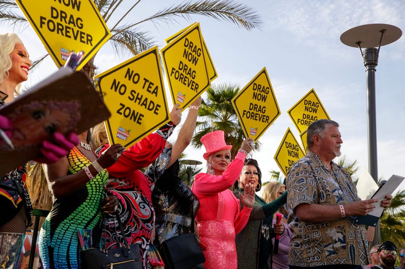 man with clipboard speaks before group of drag queens holding signs that say drag now drag forever