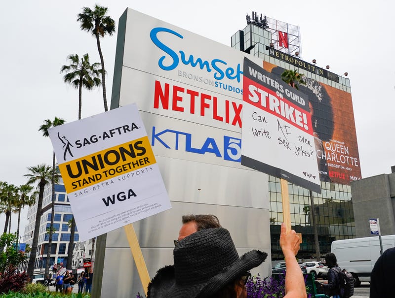 picketers with signs that say writers guild on strike in front of sunset bronson studios sign