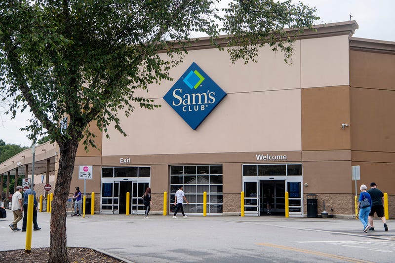 Photo of the front exterior of Sam's Club big box store with customers walking inside. A tree is in the foreground.