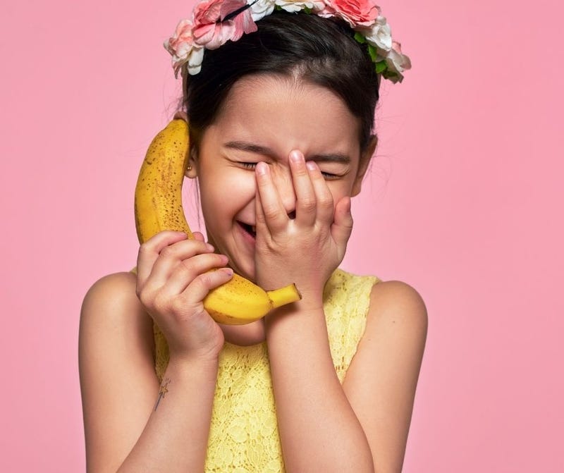 A happy kid in a yellow dress pretends to talk on a banana like by mobile phone.