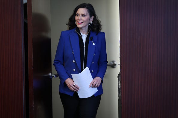 Governor Whitmer, in a blue blazer and papers in hand, enters a door looking to her right